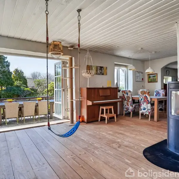 Indendørs rum med brændeovn til højre, gyngestolpe i midten, og spisebord med farverige stole i baggrunden. Døre åbner til terrasse med spisebord og udsigt til grøn have.