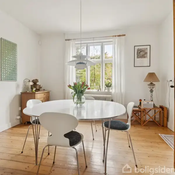 Et spisebord står i et lyst rum med trægulve. Omkring bordet er der fire stole, og på bordet står en vase med blomster. En loftslampe hænger over bordet.
