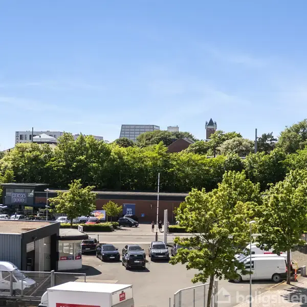 En parkeringsplads med flere biler holdende, omkranset af bygninger og grønne træer under en klar blå himmel. I baggrunden ses flere bygninger, herunder en med et tårn.