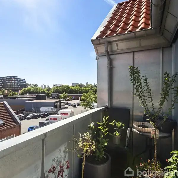 Balkonen har planter i potter og udsigt til bygninger og en parkeringsplads med køretøjer. Himmel og sollys skaber en lys atmosfære.