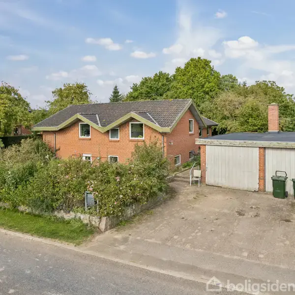 Huset er en rød murstensbygning med sadeltag, omgivet af træer og buske. En garage står til højre med to skraldespande foran. Asfalteret indkørsel leder op til huset.