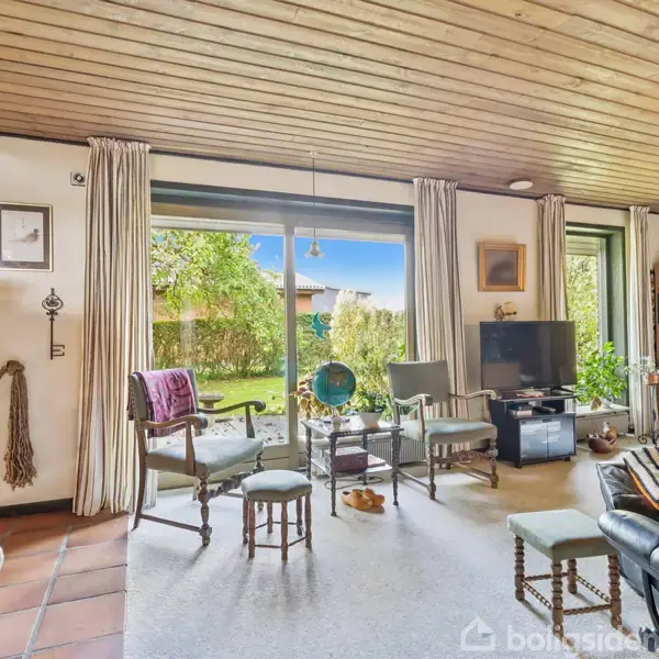 Stue med sofagruppe og fjernsyn står ved vinduer med udsigt til have. Træloft og teglstensvægge tilføjer rustik atmosfære. Bogreoler og billeder på væggen. Ingen tekst til stede.