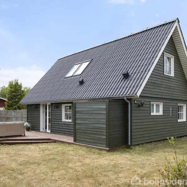 Et mørkegrønt træhus med en stejl, skråt tag ligger på en græsplæne. Foran huset er der en terrasse med udendørs sofaer. Huset er omgivet af træer.