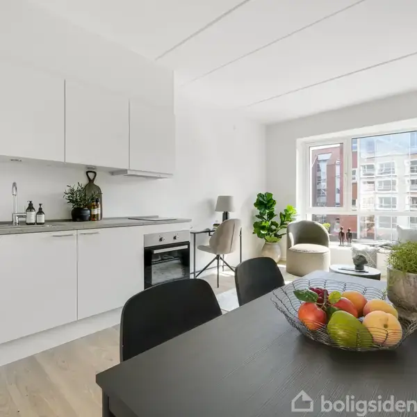Et moderne køkken-alrum med hvide skabe og en sort bordplade. Frugtskål på spisebordet. En stor vindue giver udsigt til lejligheder udenfor, og planter står i hjørnet.