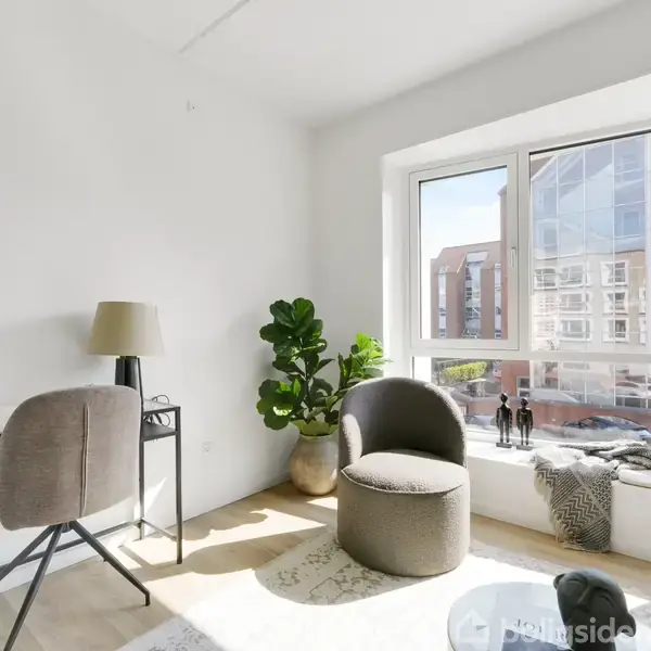Et lyst værelse indeholder en stol og et skrivebord med lampe. Ved vinduet er der en rund lænestol og en plante. Udsigt til moderne bygninger udenfor.