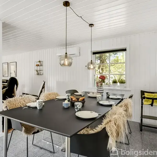 Sort spisebord dækket med tallerkener og kopper, omkranset af stole med pels. Tre hængelamper lyser rummet op. Rummeligt, lyst køkken med vindue, ur på væggen, og planter.