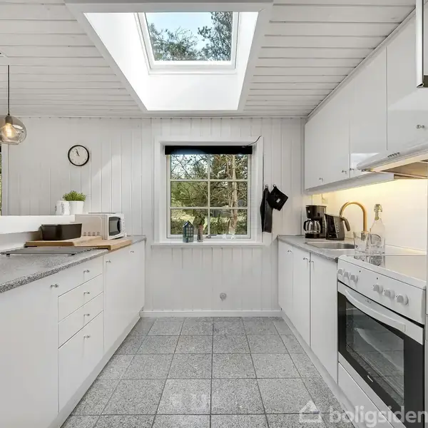 Et lyst køkken med hvide skabe og grå fliser. Et vindue og ovenlys giver naturligt lys. Køkkenredskaber står på bordpladen ved siden af en indbygget ovn og køleskab.