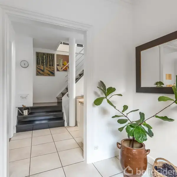 En grøn plante i en brun krukke står på gulvet. Et spejl hænger på væggen i en entre med hvide fliser, mens en trappe ses i baggrunden.