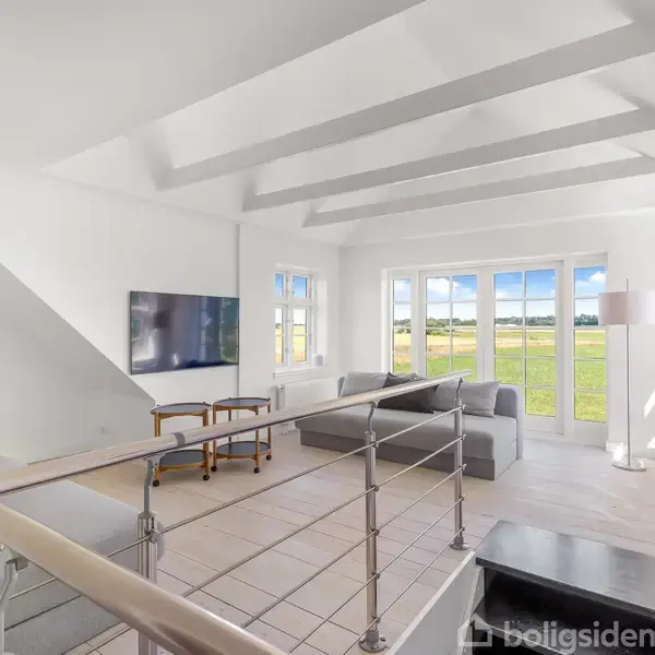 Stue med sofaer arrangeret omkring et bord, fjernsyn på væggen. Store vinduer giver udsigt til en grøn græsslette. Trærækværk og skråtstående loftstrukturer skaber moderne indtryk.