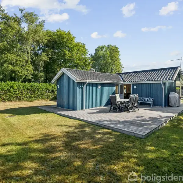 Et blåmalet sommerhus står stille på en græsplæne. Der er en stor terrasse med havemøbler. Omkring huset er der grønne træer og buske under en blå himmel.