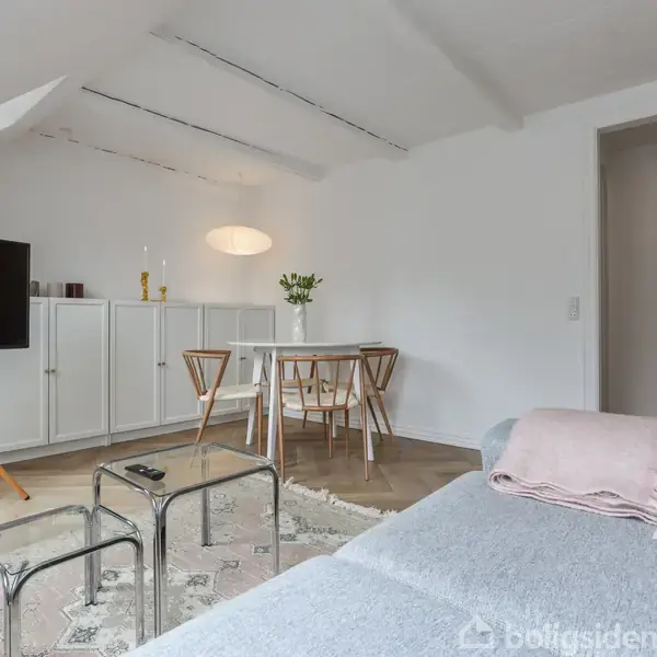 Et lyst stueområde med en grå sofa, to glasborde og et TV på stativ. Et spisebord med fire stole står i baggrunden. Rummet er minimalistisk indrettet med skråvægge.