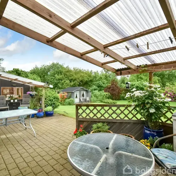 En overdækket terrasse med bord og stole, omgivet af planter og lanterner. I baggrunden ses en græsplæne med et skur og tæt beplantning, der skaber en hyggelig haveatmosfære.