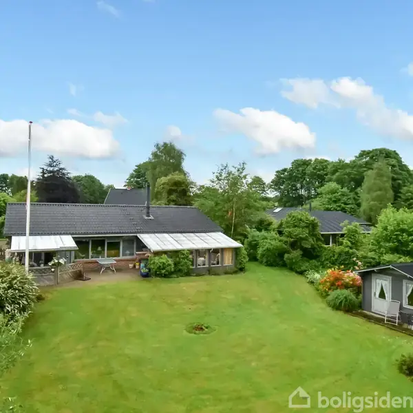 Hus med sort tag står stille i en grøn have med omgivende træer; til højre ses en lille udestue. Skyfri himmel med få skyer danner en lys baggrund.