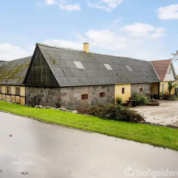 En stor, traditionel ladebygning med skrått tag står ved siden af en landevej. Græsarealer omgiver bygningen, og himlen er klar med få skyer.
