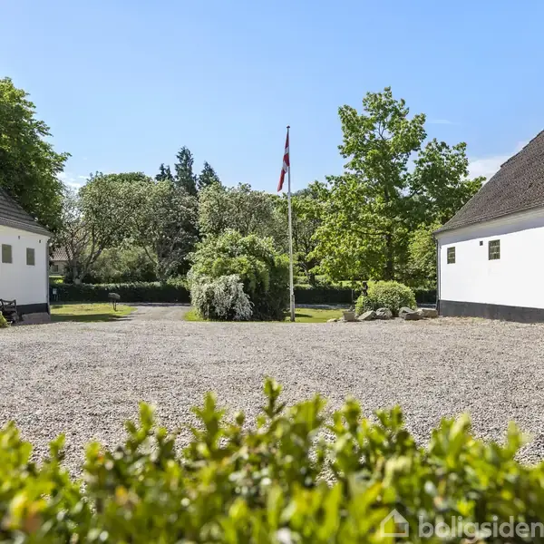 To hvide bygninger står på hver side af en grusbelagt gårdsplads. En flagstang med et dansk flag er i midten, omgivet af grønne træer og buske i baggrunden.