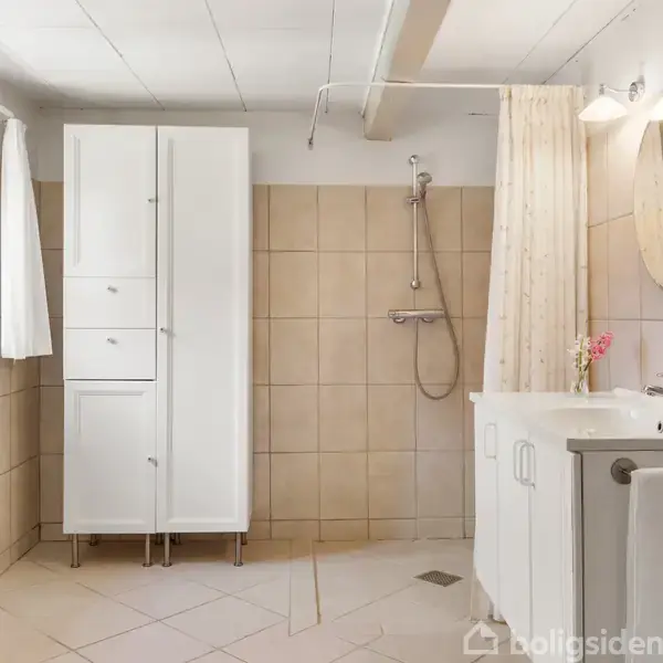Et badeværelse med hvidt skab, bruser med beige fliser, samt håndvask med rundt spejl. Miljøet er lyst med et lille vindue og hvide gardiner.