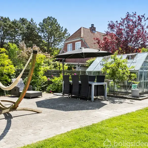 Hængekøje står ved siden af et spisebord med sorte stole og parasol. Et drivhus er placeret tæt ved, omringet af frodig vegetation og et hus i baggrunden.