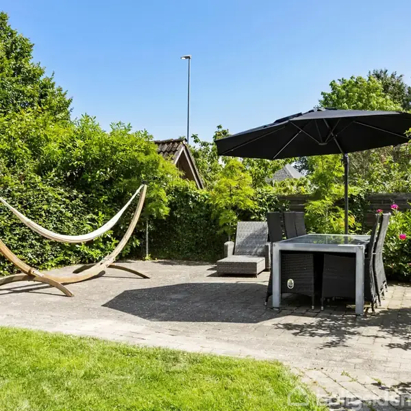 Hængekøjestativ står tomt på terrasse omgivet af frodig have. Ved siden af står et bord med stole og en stor, sort parasol. Solrig dag med blå himmel.