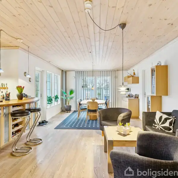 Stue med træloft og -gulv, har sofa og stole i forgrunden. En spiseplads med bord og stole ses i baggrunden ved store vinduer. Køkkenbar til venstre.