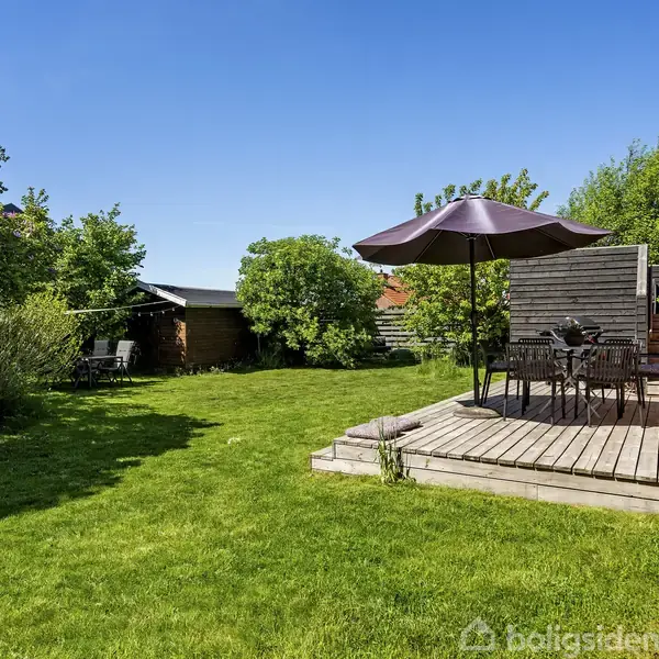 En terrasse med havebord og parasol står på trædæk ved en rød murstensbygning. Grønt græs og buske omgiver området under en klar, blå himmel.