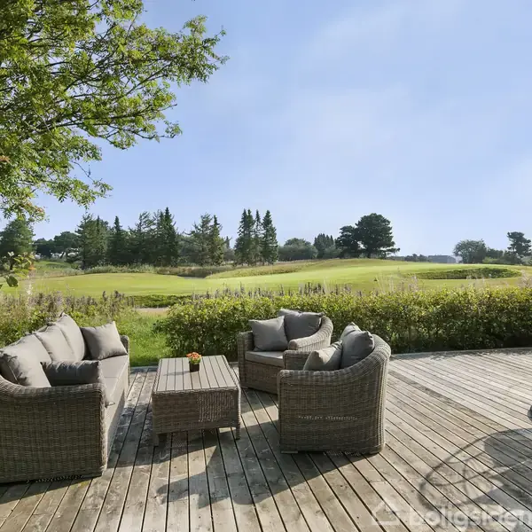 Terrassemøbler står på en træterrasse. Møblerne omfatter to sofaer og en stol omkring et lavt bord. Terrassen vender ud mod en grøn græsplæne med træer i baggrunden.