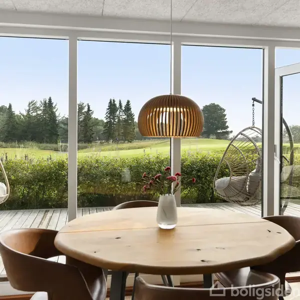 Et rundt bord med fem stole står i en spisestue med en stor glasvæg. To hængestole er udenfor på terrassen med udsigt til en græsplæne og træer.