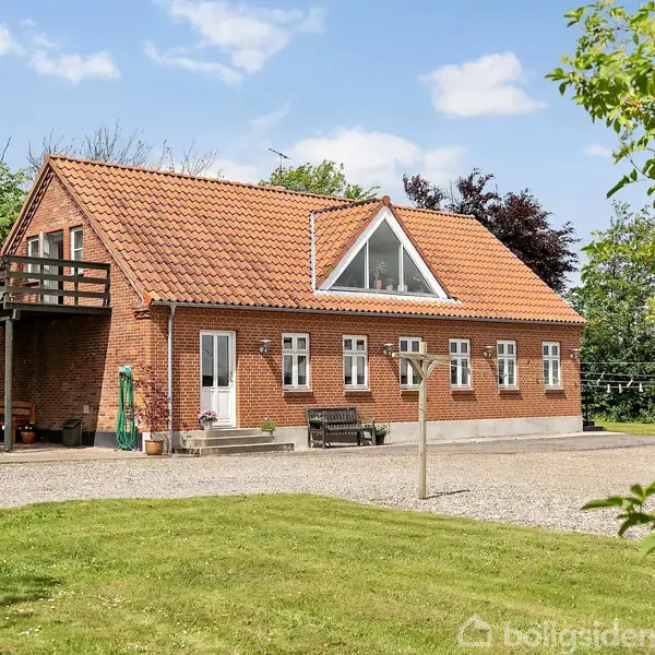 Rødt murstenshus med orange tegltag står stille i frodigt haveområde, omgivet af græsplæner og træer. En grusvej og terrasse med borde og stole danner en hyggelig atmosfære.