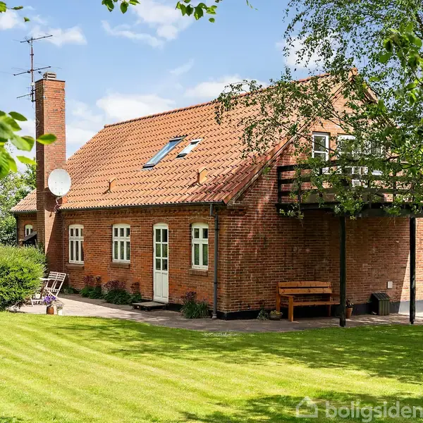 Et rødt murstenshus med skråt tegltag står i en grøn have med træer og velplejet græs. Ved huset er der en terrasse med en bænk og en satellitantenne.