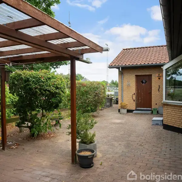 Et hyggeligt haveområde med flisebelagt gangsti, hvor en træpergola med gennemsigtig tag skaber skygge. En mindre bygning med en brun dør er i baggrunden omgivet af grønne buske.