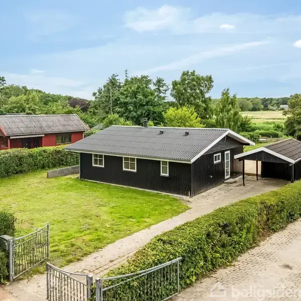 Sort træhus står stille i en grøn have omgivet af bøge. En indkørsel fører til en carport, og landskabet i baggrunden er åbent og naturligt.