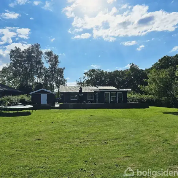 Et hus med terrasse i solskin, omgivet af en stor græsplæne og træer, under en blå himmel med skyer.