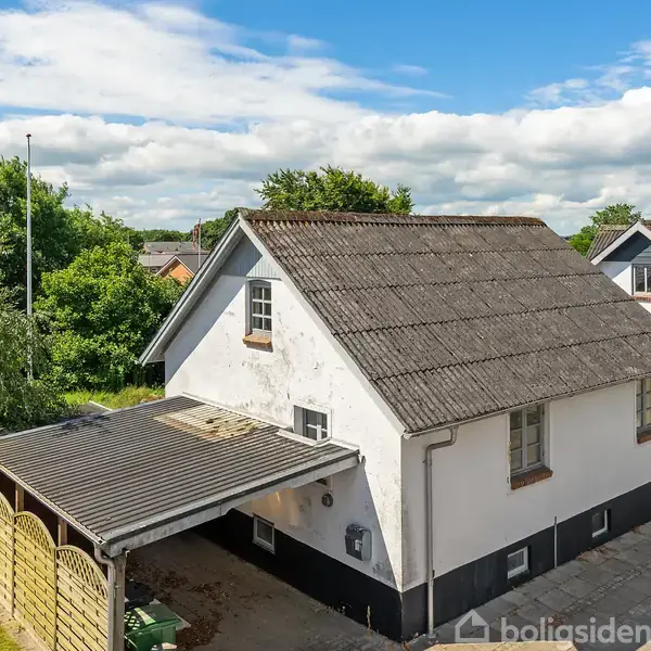 Et to-etagers hus med hvid facade og mørkt tag står stille ved siden af en carport. Græs og træer omkranser huset. Solskinsvejr med let skydække i baggrunden.
