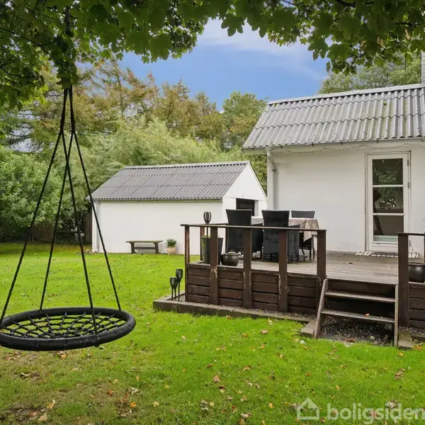 En rund gynge hænger stille fra et træ i en frodig have. Ved siden af er der en terrasse tilknyttet et hvidt hus med et bord og stole.