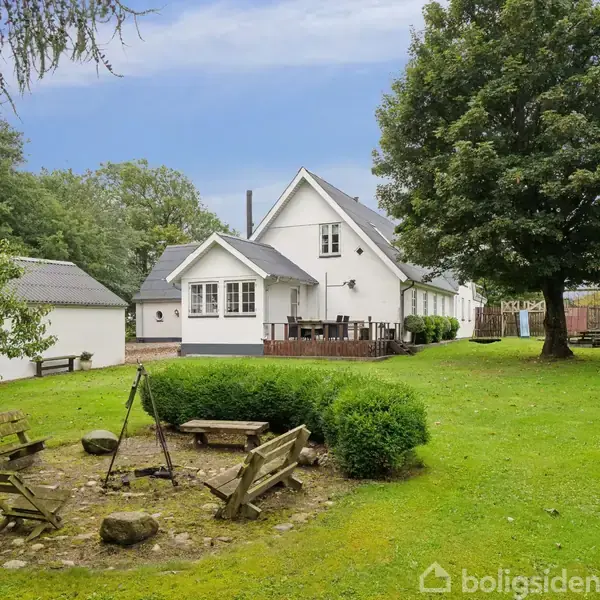 Hvidt hus med terrasse, omgivet af en grøn have. I forgrunden er en bålplads med tre træbænke og buske, omgivet af græsplæne og træer.