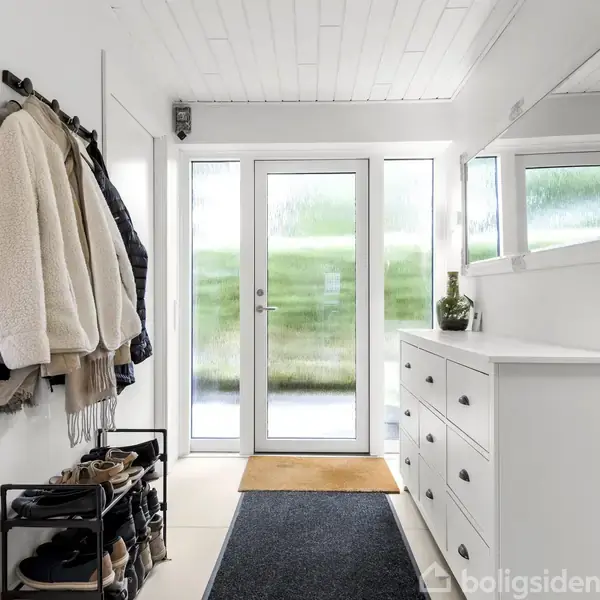 En entré med hvide vægge og loft. Jakker hænger på kroge til venstre, mens sko står på en skohylde. Til højre er en hvid kommode og et spejl. Dobbeltglasdør åbner mod en grøn have.