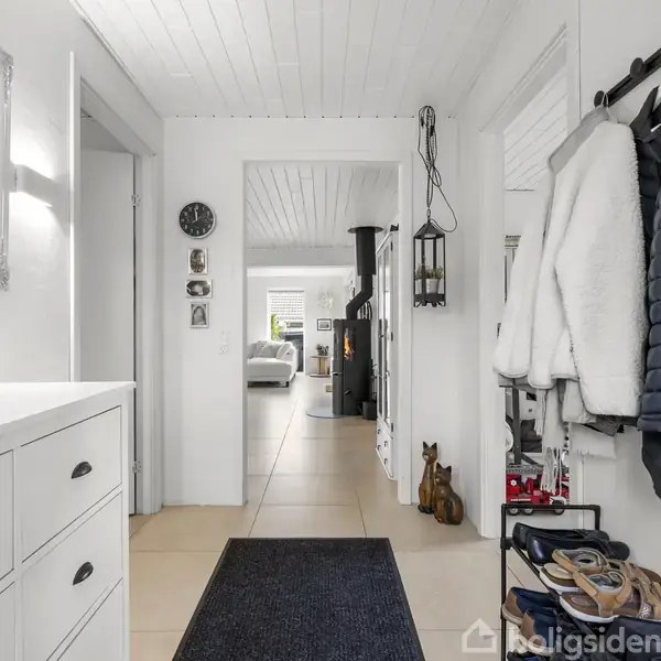 Gang med skoskab og knagerække fuld af jakker. I baggrunden ses en stue med en brændeovn og en sofa. Væggene er hvide med flere indrammede billeder og et ur.