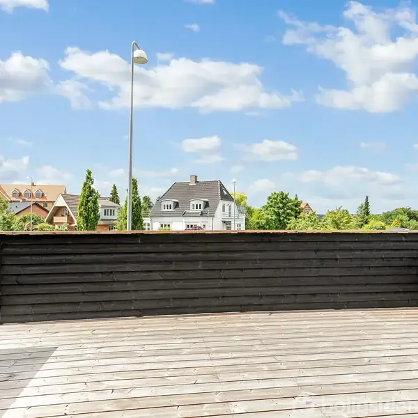 Træterrasse med sort gelænder. Set udefra med udsigt til naboers huse og en blå himmel med enkelte skyer.