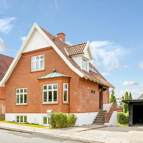 Et rødt murstenshus står stille i et boligområde med græs og fliser foran. Ved siden af er en carport med hæk. Himmelen er blå med få skyer.