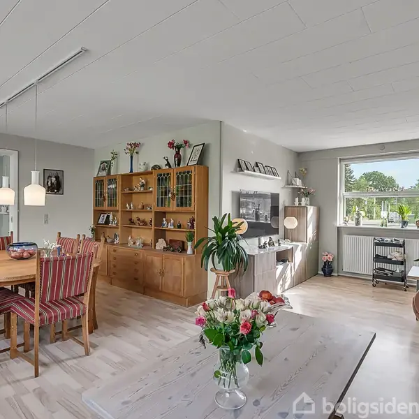 Stue med træbord og rødstole til venstre, hylder med bøger og dekorationer i midten, sofa og vindue med udsigt mod træer til højre. Blomster på bord.