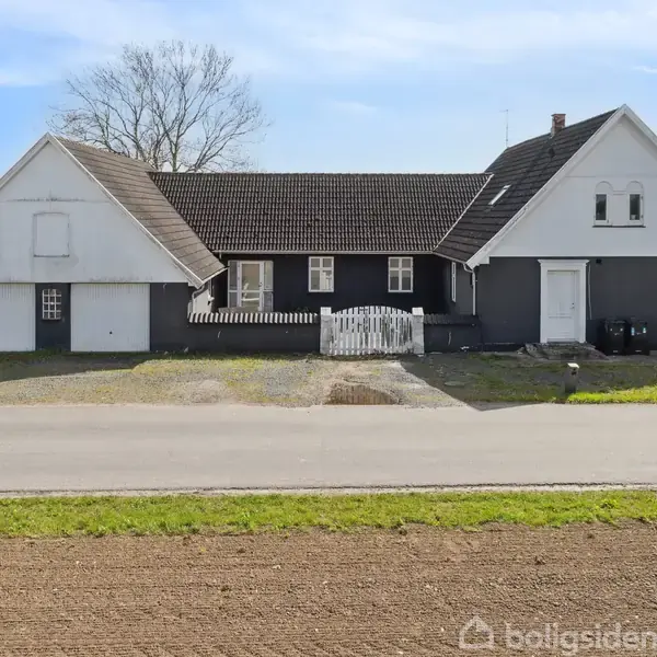 En hvid og grå husbygning med tre gavle står stille ved en vej. Den har en lille hvid havelåge og er omgivet af græs og marker.