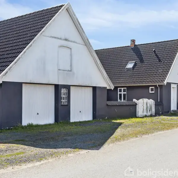 To bygninger med sorte tage og hvide døre står ved siden af hinanden på en solrig dag. De er omgivet af grus og en mindre vej.
