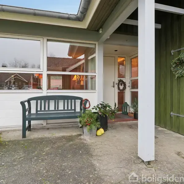 Bænk står foran husets indgang, dekoreret med planter og dørkrans. Et tag beskytter området, og vinduer viser reflektioner af nabohuse.