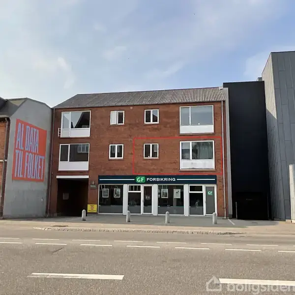 En treetagers murstensbygning med GF Forsikring nederst, placeret ved en vej. Grafitti på nabobygningen siger "AL DATA TIL FOLKET." Klare vinduer ses på facaden.