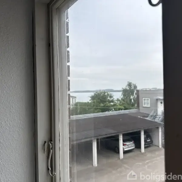 Et vindue viser en udsigt mod en carport med biler nedenfor og træer samt vand i baggrunden. Himlen er overskyet, og bygninger ses også i nærheden.