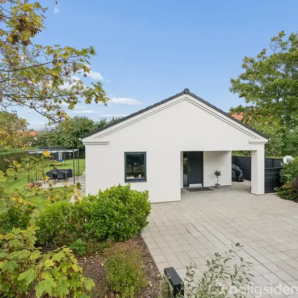 Hvidt hus med carport, omgivet af grønne træer og buske, står på en stor flisebelagt gårdsplads. En klar blå himmel skaber en lys og indbydende atmosfære.