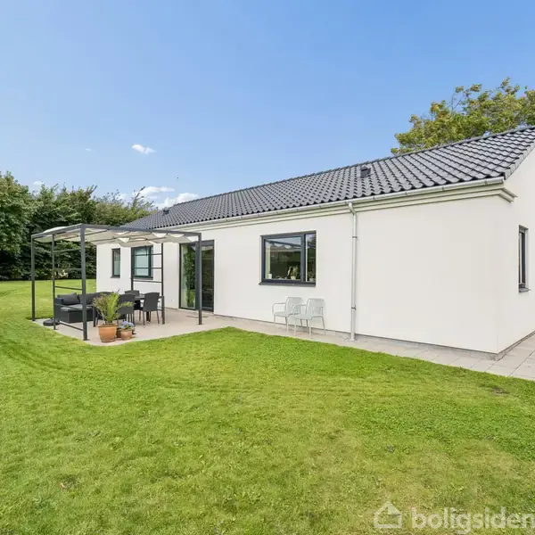 En hvid, moderne villa med sort tag står i en velplejet have med græsplæne. En terrasse med havemøbler ses til venstre, og en trampolin anes i baggrunden.
