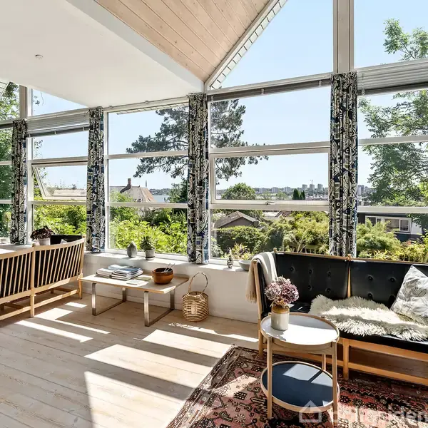 Stue med sofaer og småborde fyldt med lys fra store vinduer, omgivet af grønne planter og udendørs udsigt. Lyse trægulve og loft skaber en luftig, indbydende atmosfære.