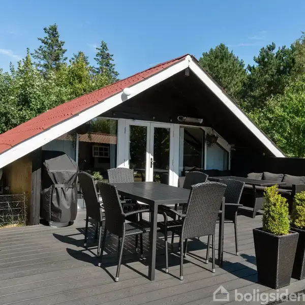 Sort udendørs terrasse med havemøbler og parasol står foran en sort hytte med rødt tag. Omgivet af grønne træer under en klar blå himmel.
