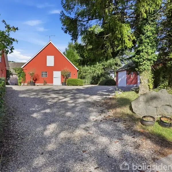 Rødt hus med gavl, omgivet af træer og grusvej, under en blå himmel. To potteplanter står ved en stor sten ved siden af vejen.