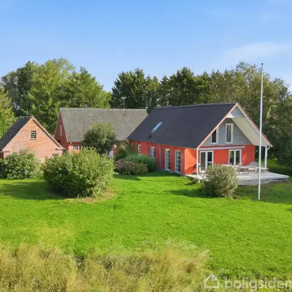 Et rødt hus med skråtag står i en grøn have omkranset af træer. En flagstang er placeret ved siden af huset. Det er solskin og himlen er klar blå.